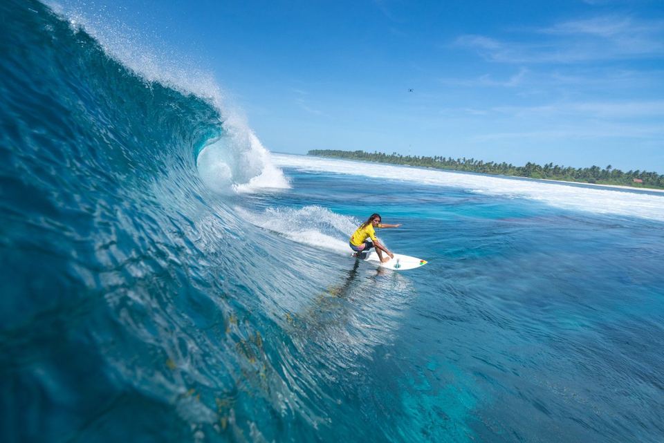 A surfer carving across a turquoise Maldivian wave beside a palm-fringed island